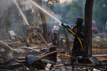 Un bombero con casco y traje oscuro rocía un chorro de agua desde una manguera amarilla sobre escombros, con niebla de agua y luces rojas difusas al fondo