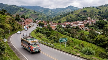 Vista aérea de una carretera en curva con un bus rojo y verde. Alrededor, montañas verdes con casas dispersas y un cartel que dice "Bienvenidos a Jardín".