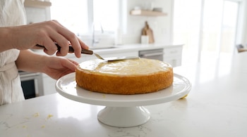 Manos de una persona aplicando glaseado con una espátula a una torta de limón sobre un pedestal blanco, con ralladura de limón visible y una cocina moderna al fondo.