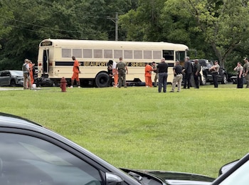 Un autobús penitenciario beige con inscripciones del sheriff, agentes policiales, y personas con uniformes naranjas en un área de césped con árboles