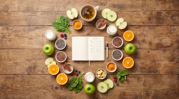 Cuaderno con un bolígrafo, manzanas verdes, naranjas, espinacas, canela, bayas, semillas de chía, semillas de lino, cúrcuma, jengibre, yogur y té en una mesa de madera.