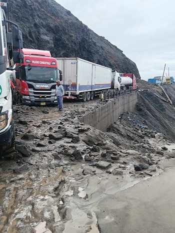 Vista de camiones detenidos en una carretera bloqueada por lodo, piedras y agua. Un hombre de pie mira hacia adelante. Una montaña rocosa se eleva a la derecha