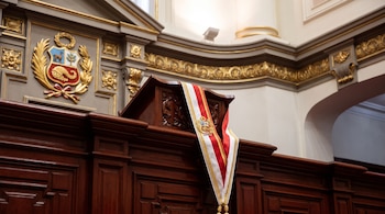 Banda presidencial roja, blanca y dorada sobre atril de madera; escudo nacional peruano, paredes con molduras doradas.