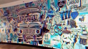 Mural colorido en un hotel, mostrando un autobús con jugadores de fútbol argentinos y el texto "Campeones del Mundo", con un efecto visual distorsionado