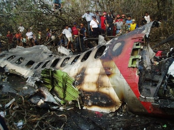 Restos destruidos y calcinados del fuselaje de un avión rodeados por personas en un terreno con árboles y ramas