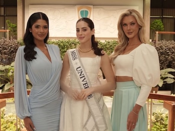 Tres mujeres jóvenes con vestidos elegantes posan de pie en un interior; una lleva un vestido azul, otra un vestido blanco con banda "Miss Universe", y la tercera un top blanco con falda turquesa