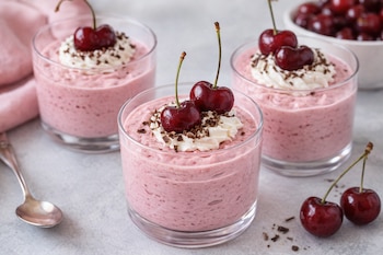 Tres vasos de mousse de cereza con nata montada, virutas de chocolate y cerezas frescas encima, junto a una cuchara y un paño rosa.