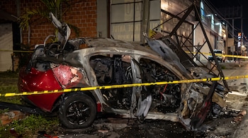 Automóvil rojo calcinado y deformado tras un impacto, rodeado por cinta amarilla de precaución policial en una calle nocturna.