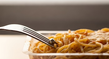 Primer plano de un tenedor metálico que se inserta en un plato de espagueti horneado en bandeja plástica, con un fragmento de metal visible entre la pasta.
