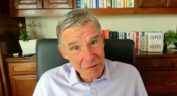 Hombre de mediana edad con camisa sentando en escritorio de madera. Fondo con estanterías, libros variados y plantas. Libro 'Super Agers' visible