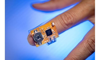 Primer plano de un dedo sosteniendo un circuito electrónico flexible naranja con microchip, batería plateada y otros componentes. Fondo azul