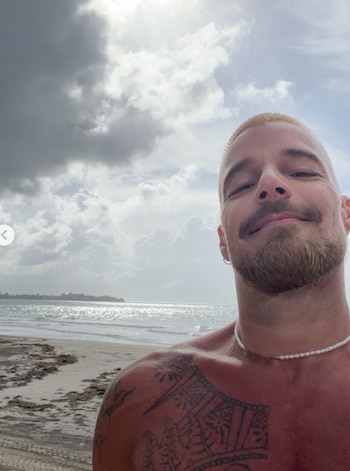 Hombre sin camiseta en traje de baño saltando en la arena de una playa, con el mar, rocas, árboles y cielo nublado