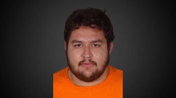 Retrato de un hombre joven con cabello oscuro y rizado, barba, bigote y camiseta naranja, mirando al frente, sobre un fondo degradado gris oscuro.