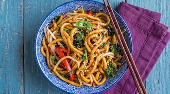 Cuenco azul con fideos, brotes de soja, pimiento rojo, hojas verdes, semillas de sésamo, cilantro. Palillos de madera y una servilleta púrpura en mesa azul.