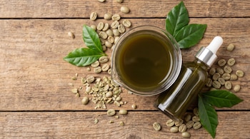 Una tabla de madera con granos de café verde, hojas, un vaso de precipitados con líquido verde y una botella de gotero con líquido verde.