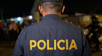 Vista de la espalda de un hombre con uniforme azul oscuro y gorra. La palabra "POLICIA" bordada en letras amarillas se observa en su espalda. Fondo oscuro y borroso.