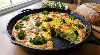 Frittata de brócoli, cebolla y queso en sartén de hierro fundido, una porción cortada, ensalada verde en tazón blanco, pan de hogaza y mesa de madera.