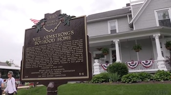 Un marcador histórico marrón con texto y bandera de Estados Unidos delante de una casa gris de dos pisos con porche blanco, adornos patrióticos y césped