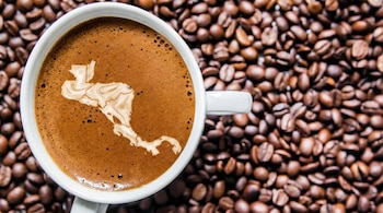 Una taza blanca de café oscuro con el mapa de Centroamérica delineado en espuma clara, rodeada por una superficie cubierta de granos de café tostado.