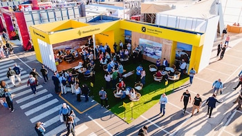 Vista aérea de un evento al aire libre. Stands amarillos, césped artificial, zonas de descanso y multitudes de personas caminando e interactuando