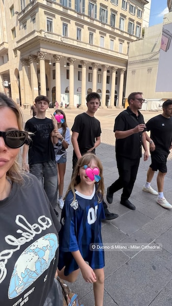 Primer plano de una mujer con gafas de sol, junto a varios niños y adultos caminando por una calle. Fondo con edificio de columnas. Etiqueta 'Duomo di Milano'