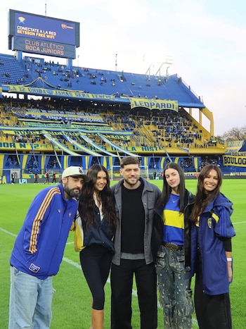 Cinco personas sonríen en el campo de fútbol verde de un estadio. Las gradas azul y amarillo están llenas de aficionados. Hay una pantalla gigante al fondo