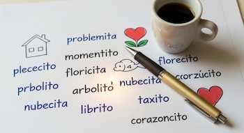 Una hoja de papel con palabras escritas en diminutivo y dibujos de una casa, un corazón y una nube, junto a un bolígrafo, una taza de café y una pequeña llama.
