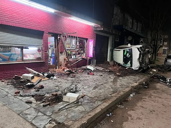 Fachada de ladrillo rojo de un almacén destruida por impacto, con escombros y vidrios en la acera; una camioneta blanca volcada sobre su lateral