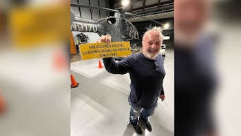 Un hombre con barba sostiene un cartel amarillo frente a un helicóptero negro dentro de un hangar