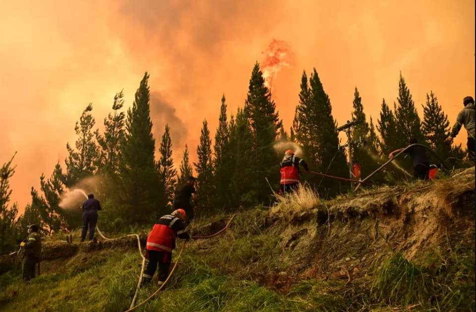 Un gran incendio forestal en Chubut emite intensas llamas y humo naranja bajo la luna, mientras vehículos de emergencia se congregan en una carretera cercana listos para actuar (Imagen Ilustrativa Infobae)