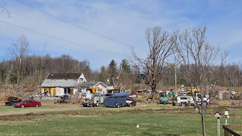 Paisaje rural tras desastre con casas dañadas, árboles quebrados, escombros, un coche rojo, varios camiones de trabajo y una excavadora verde en un campo