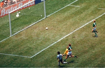 Cancha de fútbol con césped, arco blanco y pelota. Tres jugadores en acción: uno con camiseta celeste y blanca, dos con camiseta verde. Público en tribunas
