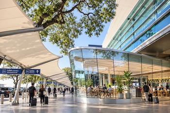 Personas con maletas caminan bajo una cubierta exterior junto a un edificio moderno de cristal con mesas, sillas y señales de salidas y arribos