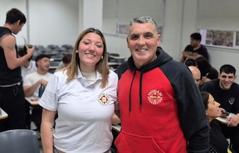 Un hombre y una mujer joven sonríen de pie en un aula mientras llevan indumentaria con el escudo de la Escuela de Guardavidas