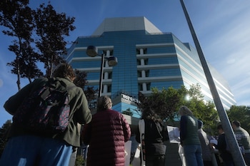 Vista desde atrás de un grupo de personas frente a un edificio moderno con fachada de cristal azul y blanco, bajo un cielo azul claro. Hay árboles y un poste de luz