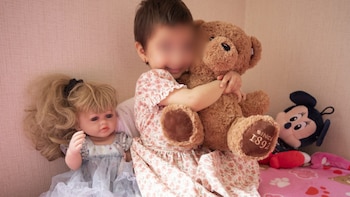 Niña sentada abraza un oso de peluche, junto a una muñeca de cabello rubio y una figura de Minnie Mouse