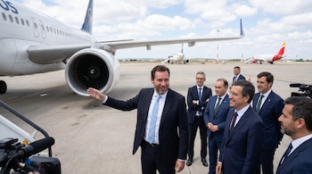 Oscar Puente, un hombre de traje azul y corbata clara, señala un avión blanco grande en la pista de un aeropuerto, acompañado por otros hombres de traje.