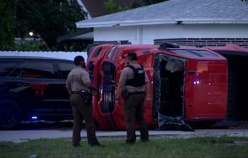 Un SUV rojo volcado con ventana rota yace al lado de una camioneta policial negra K-9, con dos oficiales uniformados observando el lugar del choque