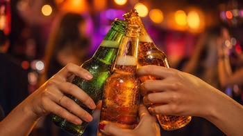 Tres manos sostienen y chocan botellas de cerveza de vidrio verde y ámbar ante un fondo con luces desenfocadas