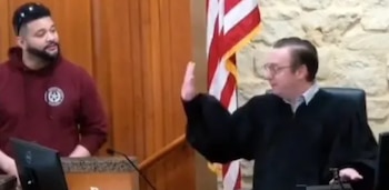 Nathan Milliron, con sudadera oscura, de pie junto a un juez en toga que levanta la mano, con una bandera de EE. UU. y pared de piedra de fondo en un tribunal