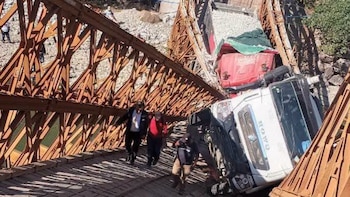 Un puente metálico colapsado con dos camiones de carga atrapados entre la estructura inclinada y tres personas caminando por el piso de madera