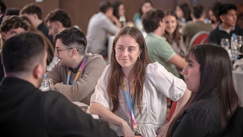 Una joven con blusa blanca y credencial al cuello conversa con personas sentadas en una mesa dentro de un salón concurrido
