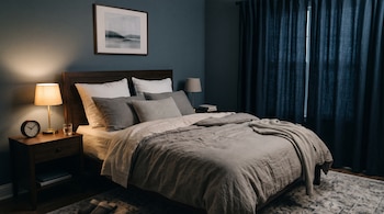 Cama matrimonial con sábanas grises, mesa de noche con lámpara encendida y reloj, cuadro en la pared y cortinas oscuras cerradas.