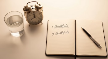 Vista cenital de una mesa de luz blanca con un vaso de agua, un despertador dorado y un cuaderno abierto con la palabra 'Gratkfuls' escrita, junto a un bolígrafo.