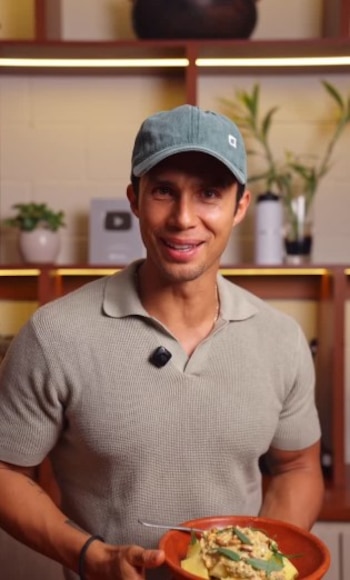 Rodrigo Fernandini sonríe a cámara, con gorra verde y polo, sosteniendo un plato de pasta con salsa clara, hojas verdes y frutos secos. Al fondo, estantes con objetos decorativos