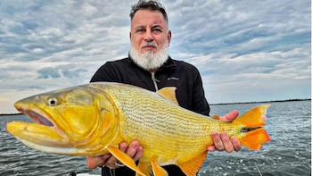 Un hombre con barba canosa sostiene un pez dorado grande frente a un cuerpo de agua bajo un cielo nublado