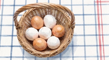 Una cesta de mimbre con siete huevos, cuatro blancos y tres marrones, sobre un mantel blanco con patrón de cuadros azules y rojos, en estilo de acuarela.