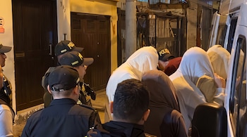 Policías con gorras oscuras conducen a varias personas cubiertas con capuchas blancas hacia un vehículo blanco de noche.