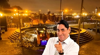 Un hombre sonriente en camisa blanca y corbata apunta a la cámara, con una máquina excavadora removiendo escombros en el fondo, bajo luces amarillas nocturnas.