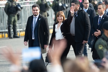 Una mujer de cabello castaño, chaqueta oscura y blusa blanca sonríe y saluda, acompañada de hombres en traje, con personal de seguridad al fondo